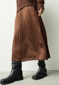 Hnedá plisovaná sukňa s plynulou textúrou, skombinovaná s čiernymi koženými čižmami po kolená s detailom remienka a pracky.