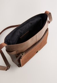 Sac bandoulière en cuir marron avec poche avant zippée, intérieur en tissu noir et bandoulière réglable sur un fond clair.