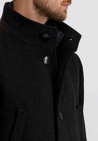 Schwarzer Wollmantel mit hohem Kragen, zwei Fronttaschen und schwarzen Knöpfen. Verfügt über einen strukturierten Stoff und einen Reißverschluss am Hals.