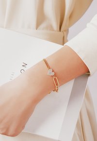 Guldarmband med ett hjärtcharm prytt med rhinestones och ett pappersklipsaccent, bärs på en handled, mot en ljus bakgrund.