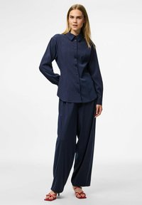 Jūras spēku svītrainā krekls ar pogām un apkakli, apvienots ar vaļīgām jūras spēku biksēm; audumam ir gluda tekstūra un smalka spīdība.