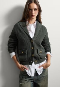 Cardigan verde a coste con bottoni dorati e tasche frontali, indossato sopra una camicia bianca. Tessuto a maglia testurizzato con design a collo sciallato.
