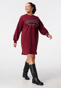 Bordeaux sweatshirt jurk met lange mouwen, voorzien van de tekst "BROOKLYN" en zijsplitten. Gecombineerd met kniehoog zwarte leren laarzen.