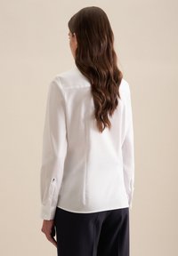 Blusa bianca a maniche lunghe con colletto, texture liscia, polsini con bottoni, spacchi laterali e vestibilità tailored, vista da dietro.