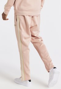 Světle růžové teplákové kalhoty z hladkého materiálu, s bílými a zlatými bočními pruhy, elastickým pasem a volným střihem.