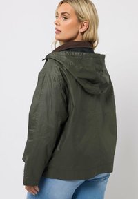 Veste à capuche vert olive avec un col en cuir texturé marron, présentant une coupe ample et des détails de couture visibles.