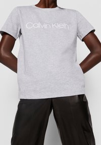 Světle šedé bavlněné tričko s kulatým výstřihem a krátkými rukávy, se bílým logem "Calvin Klein" napříč přední stranou. V kombinaci s černými kalhotami.