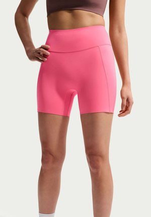 Kobieta ubrana w jasnoróżowe, sportowe szorty z wysokim stanem i mauve stanik sportowy, stojąca z jedną ręką na biodrze na jednolitym tle.