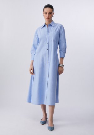 Femme portant une robe chemise midi bleu clair à boutons, manches trois-quarts et ballerines pointues bleues devant un fond uni.