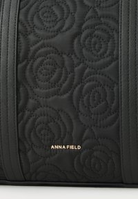 Quiltana crna torba sa uzorcima ruža, istaknuta glatkim tamnim trakama i zlatnim logotipom "ANNA FIELD". Izdržljiv materijal.