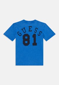 Lyseblå t-skjorte med "GUESS" i fete svarte bokstaver og "81" under, vist mot en enkel hvit bakgrunn.