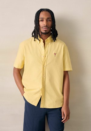 Muž s dredmi, ktorý má na sebe žltú krátku košeľu s malou vyšívanou kokteilovou pohárom a tmavé nohavice, stojí pri béžovej stene.