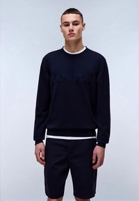 Mlad moški v črni dolg rokav sweatshirtu in črnih kratkih hlačah, ki stoji ob enobarvni svetli ozadju.