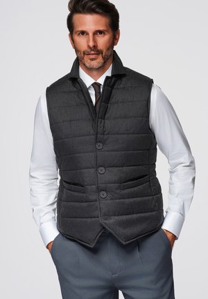 Mand iført mørkegrå quiltet knappet vest over hvid skjorte og mønstret slips, hænderne i lommerne, foran ensfarvet baggrund.