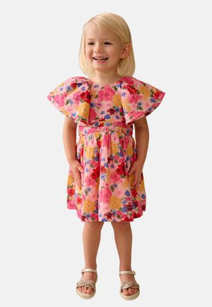 Uśmiechnięta dziewczyna o blond włosach, ubrana w różową sukienkę w kwiaty z dużymi falbanami na rękawach i złote sandały, na białym tle.