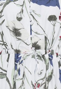 Tissu blanc avec imprimé floral vert et noir, comprenant de petits boutons blancs et une ceinture à nouer à la taille.