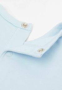 Světle modrý bavlněný oděv s detailním pohledem na složený límec s kovovými patentkami a žebrovanou texturou.