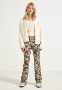 Witte, fluffy cardigan over een beige top, gecombineerd met leopardprint flared broeken. Casual sneakers maken de outfit compleet. Lichte achtergrond.