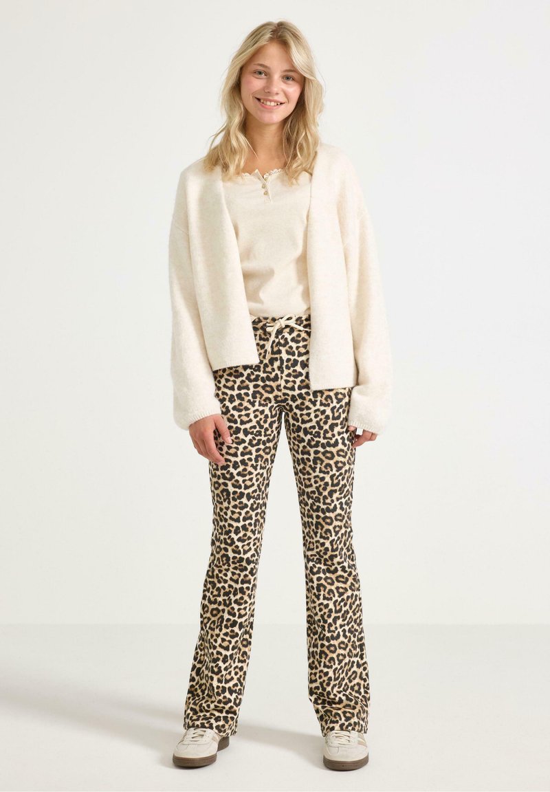 Witte, fluffy cardigan over een beige top, gecombineerd met leopardprint flared broeken. Casual sneakers maken de outfit compleet. Lichte achtergrond.