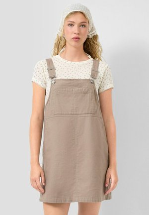 Młoda kobieta o jasnych kręconych włosach, ubrana w białą koszulę w wzory, beżową sukienkę na szelkach oraz biały chustę z koronkową lamówką, na jednolitym tle.