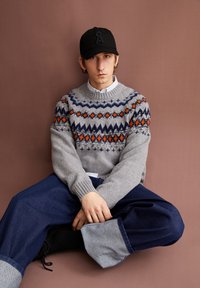 Grauer gestrickter Pullover mit mehrfarbigen geometrischen Mustern, kombiniert mit lockeren blauen Jeanshosen mit grauen Bündchen. Eine schwarze Kappe rundet das Outfit ab.