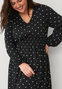 Schwarzes Polka-Dot-Kleid mit V-Ausschnitt, gerafftem Bund und langen Ärmeln. Hergestellt aus leichtem Stoff mit weißen, runden Mustern.