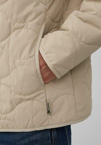Ruka v béžovej prešívanej bunde s okrúhlym prešitím a viditeľnou nášivkou "Softshell Casual" pri leme.