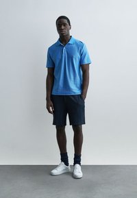 Muž stojí před jednoduchým světlým pozadím, má na sobě modré polotriko s vzorem, tmavé šortky, tmavé ponožky a bílé tenisky.