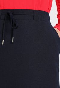 Gros plan sur une jupe marine avec taille élastique à cordon et un haut à manches longues rouge, une main glissée dans la poche de la jupe côté droit.