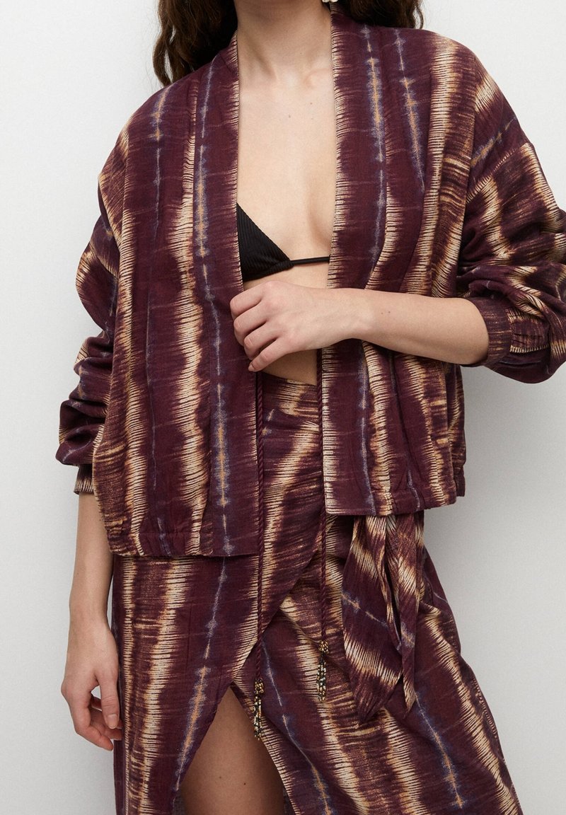 Femme portant une veste tie-dye bordeaux et beige et une jupe assortie, affichant un haut de bikini noir en dessous, avec une main proche de sa poitrine.