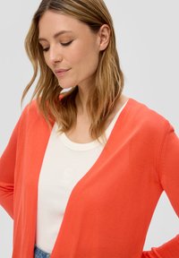 Cardigan lungo color corallo realizzato in un tessuto a maglia leggero, con scollatura a V e apertura frontale. Sotto, è visibile un top crema.