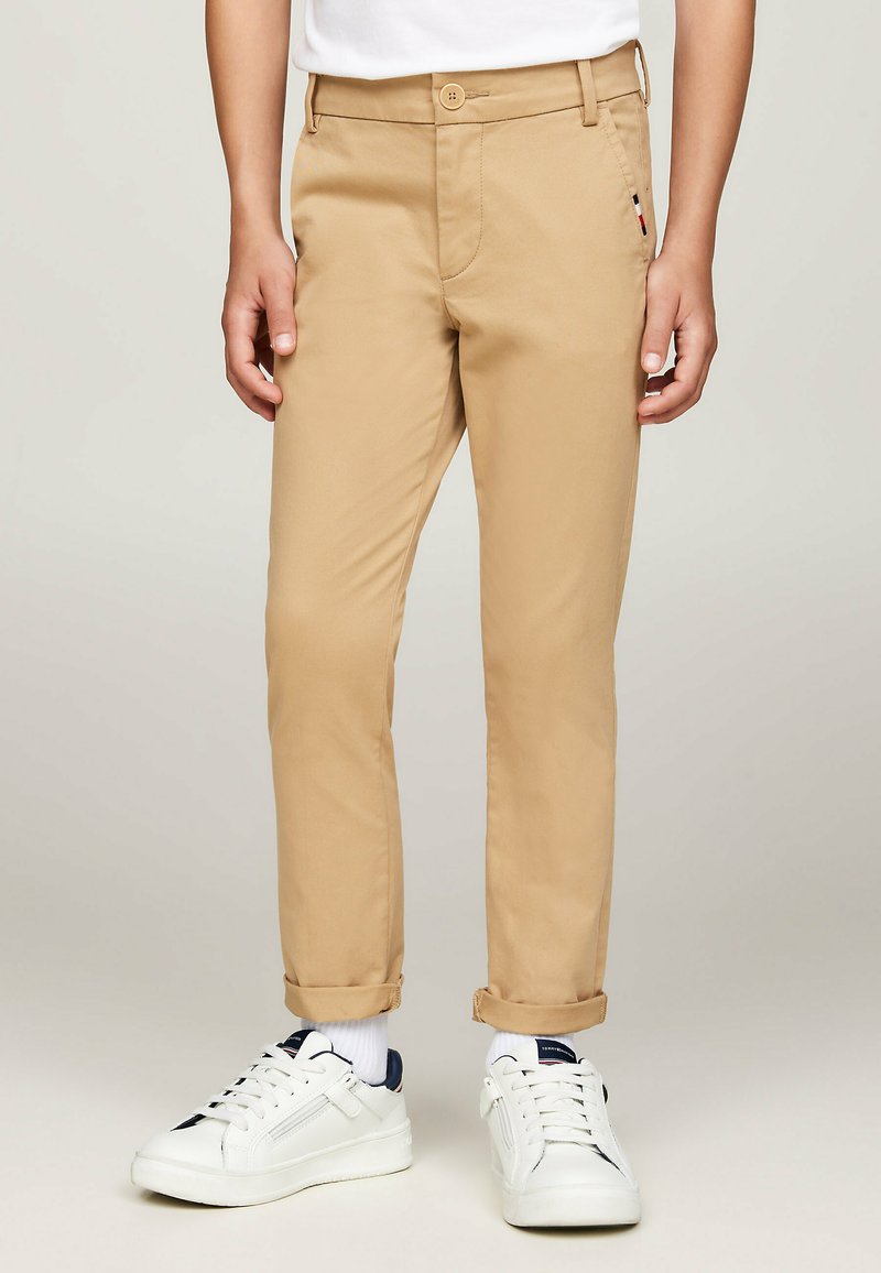 Chinos beige con una vestibilità slim, dotati di chiusura a bottoni, tasche laterali e orli arrotolati, abbinati a sneakers bianche e calzini bianchi.