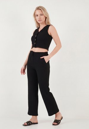 Blonde Frau steht mit einer Hand in der Tasche, trägt ein schwarzes ärmelloses, kurzes Top, passende Hose und schwarze Slip-On-Sandalen.