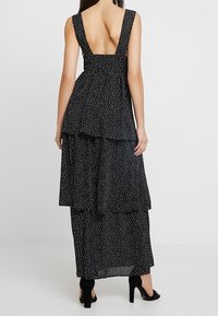 Robe noire à volants avec un motif à pois, dos en V profond, larges bretelles et silhouette fluide. Tissu texturé avec un design en couches.