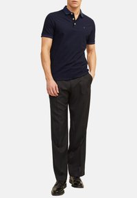 Marineblauwe polo met contrasterende kraag, drie-knopen sluiting en klein logo. Gecombineerd met zwarte broek en zwarte schoenen.