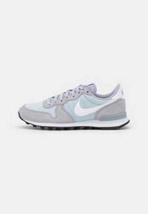 Lysegrå og blegblå Nike-sneaker med hvid swoosh, grå ruskindsoverlejringer og sort sål vist i siddeprofil på hvid baggrund.