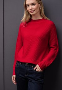 Czerwony sweter z prążkowanej dzianiny, z okrągłym dekoltem i długimi rękawami, zestawiony z ciemnoniebieskimi dżinsami. Materiał ma teksturowany wygląd.
