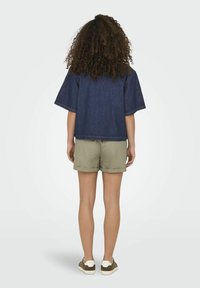 Camicetta in denim con maniche corte e larghe, abbinata a pantaloni corti color khaki chiaro. Presenta una texture di tessuto naturale e una vestibilità casual. Sneakers con suola bianca.