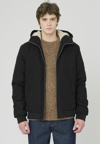 Veste zippée noire avec capuche, dotée d'une doublure beige douce et de poignets côtelés. Portée par-dessus un pull en tricot marron et un jean bleu.