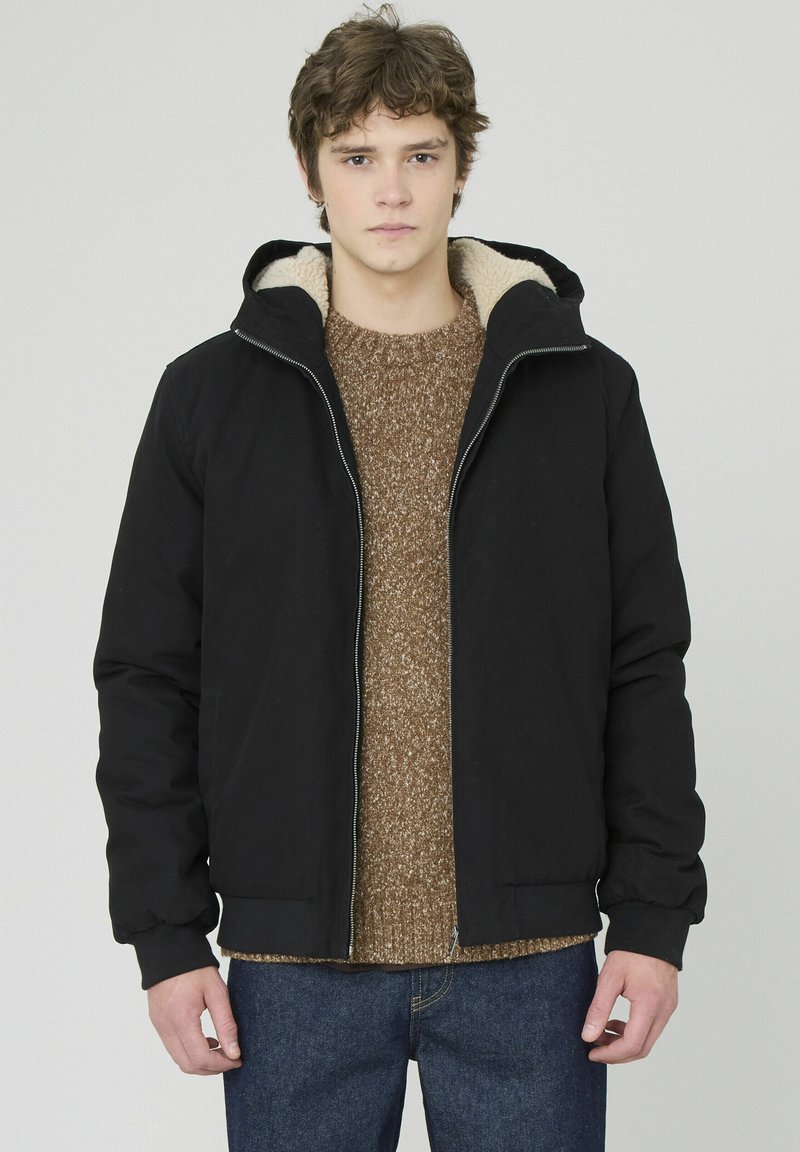 Veste zippée noire avec capuche, dotée d'une doublure beige douce et de poignets côtelés. Portée par-dessus un pull en tricot marron et un jean bleu.