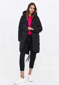 Czarna kurtka puchowa z kapturem, o długim, luźnym kroju i diagonalnym wzorze pikowania. Nosi ją osoba w różowej bluzie i czarnych spodniach.