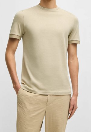 Mand iført beige kortærmet tekstureret striktrøje og matchende teksturerede beige bukser, står med den ene hånd i lommen.