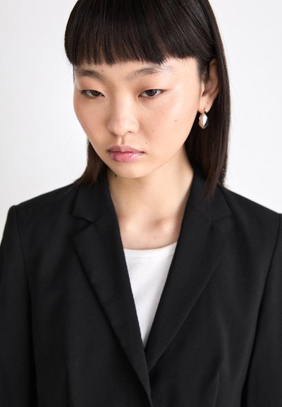 Blazer noir sur un haut blanc, avec un col en V. Le mannequin a les cheveux noirs raides et porte des boucles d'oreilles géométriques en argent.
