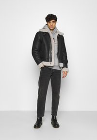 Schwarze Lederbomberjacke mit einem Schaffellkragen, kombiniert mit einem grauen Hoodie und dunklen Jeans. Schwarze Stiefeletten runden das Outfit ab.