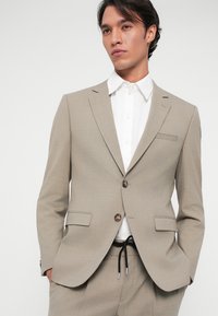 Vaaleanbeige, tekstuuroitu bleiseri, jossa on kaksi nappia ja taskut, päällä valkoinen paita. Huomionarvoisia yksityiskohtia ovat slim fit -leikkaus ja moderni muotoilu.