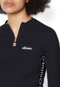 Black zip-up top with a round neckline, featuring a textured logo patch on the chest and patterned side accents. Made from a smooth fabric.