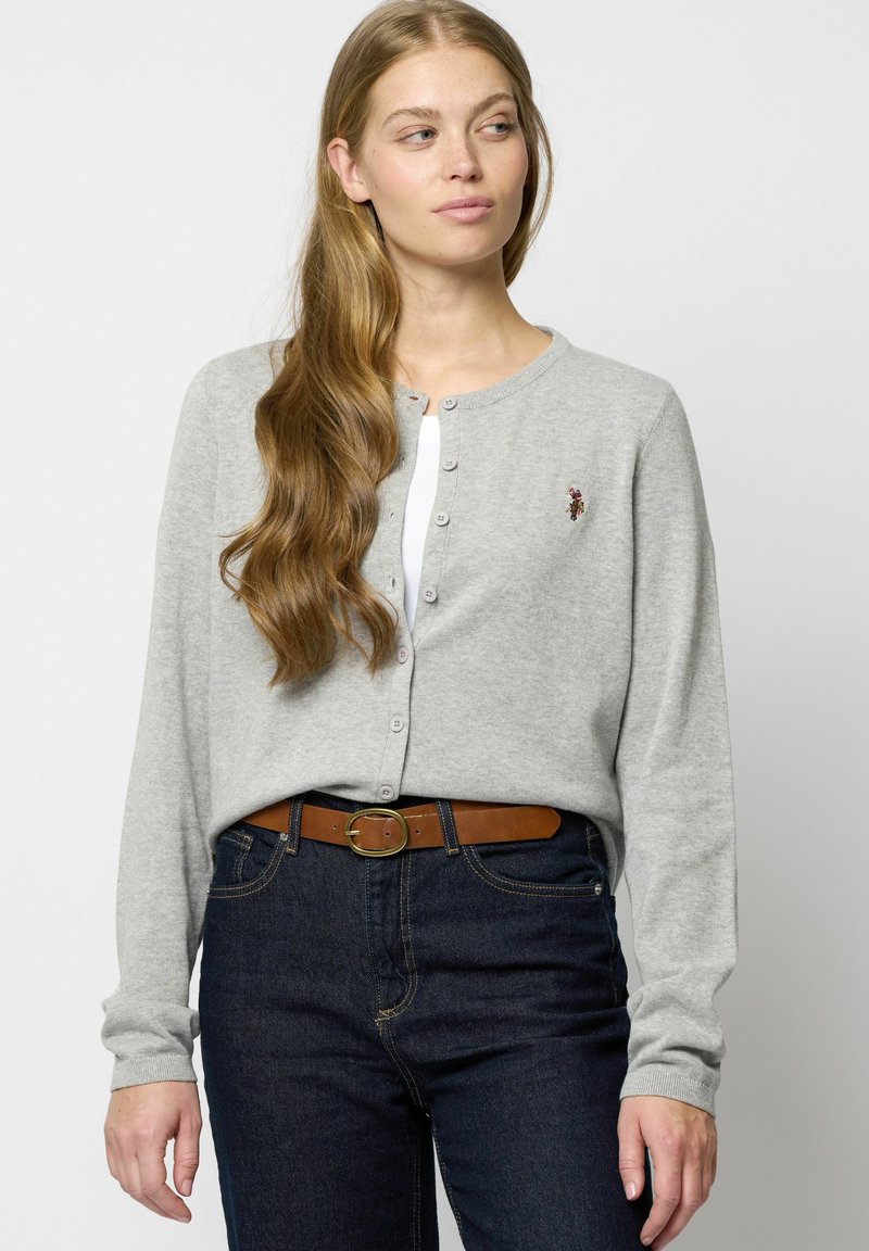 Grijze gebreide cardigan met knoopsluiting, lange mouwen en een klein geborduurd detail. In combinatie met donkerdenim jeans en een bruine riem.