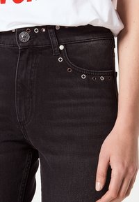 Gros plan sur un jean en denim noir avec des œillets métalliques le long de la poche avant et de la ceinture, porté avec une chemise blanche et une main visible.