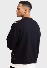Schwarzer Strickpullover mit Rundhalsausschnitt, der orange und weiße Akzente an den Schultern sowie seitliche Details mit Reißverschluss aufweist. Lässige Passform.