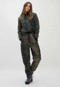 Frau steht mit den Händen in den Taschen, trägt eine gesteppte Jacke mit Leopardenmuster und passende Hose, dazu dunkle Wanderschuhe, vor einem schlichten Hintergrund.